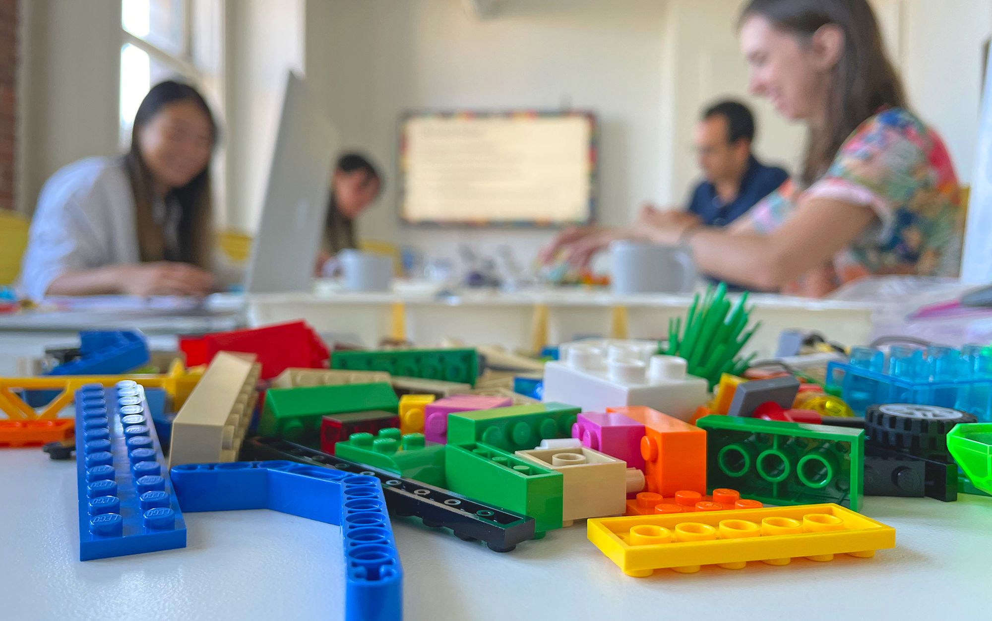 A pile of colourful assorted LEGO bricks scattered on a table in the foreground with four workshop participants blurred in the background building their own models in a boardroom